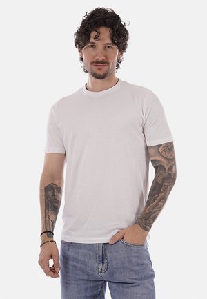 Giovane uomo con capelli ricci, che indossa una maglietta bianca semplice e jeans blu, mostrando tatuaggi dettagliati su entrambi gli avambracci.