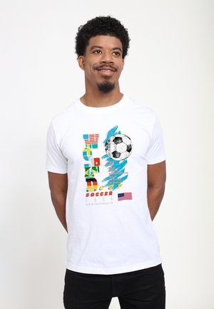 Homme portant un T-shirt blanc avec un graphisme coloré de la Coupe du Monde de football 1994, un ballon de football et le drapeau des États-Unis, debout devant un fond blanc uni.