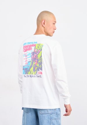 Mand iført hvid langærmet T-shirt med farverig bjergbestigergrafik og tekst på ryggen, parret med lyseblå denimshorts.