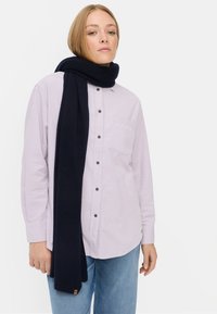 Eine marineblaue Schal um den Hals, kombiniert mit einem helllila Hemd mit Knöpfen und hellblauen Jeans. Der Schal hat eine weiche Textur.