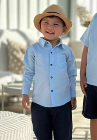 Ljusblå långärmad skjorta med knapp-ner krage, bröstficka och mörka knappar, tillsammans med marinblå shorts. Barn som bär en halmhat.