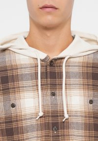 Chemise à capuche à carreaux bruns avec une capuche crème, dotée de boutons, de textures contrastées et d'un mélange de tons clairs et foncés dans le motif.