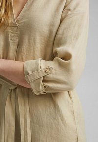 Personne portant une chemise beige à manches longues avec les manches retroussées maintenues par une lanière en tissu boutonnée.