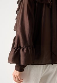 Brun, gennemsigtig bluse med flæsedetaljer langs ærmerne og en samlet manchet. Har en knaplukning ved håndleddet.
