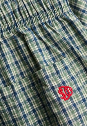 Tissu à carreaux verts, bleus et blancs avec une ceinture élastique et un petit logo rouge brodé sur une poche.