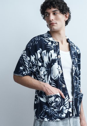 Young man with curly hair wearing a black and white floral button-up shirt over a white tank top, hand in shirt pocket, looking sideways.