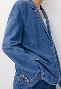 Blue denim blazer with a notched collar, buttoned cuffs, and a relaxed fit. Features visible stitching and side pockets.
