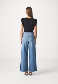 Svart topp med volangärmar, ihop med högmidjade, vida blå jeans. Tyget verkar mjukt med en slät yta.