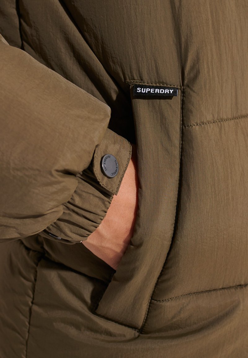 Chaqueta acolchada verde oliva con puños acanalados y una prominente etiqueta negra de Superdry en la manga. Presenta un tejido suave y texturizado.