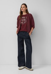 Burgunderfarbene Zip-Jacke über einem kastanienbraunen Grafik-T-Shirt, kombiniert mit dunklen, weiten Jeans und beige Sneakers, vor einem schlichten grauen Hintergrund.