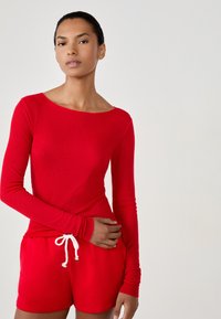 Czerwona bluzka z długim rękawem i prążkowaną fakturą o szerokim dekolcie, w zestawie z czerwonymi szortami z białym sznurkiem. Gładka tekstura, dopasowany krój.