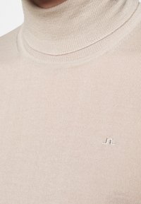 Beżowy sweter z golfem, charakteryzujący się prążkowaną teksturą oraz małym haftowanym logo z przodu. Wykonany z miękkiego, elastycznego materiału.