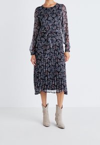 Långärmad, marinblå klänning med blommönster, midjeband och lager på lager-textur. Bärs med ljusgrå cowboystövlar.