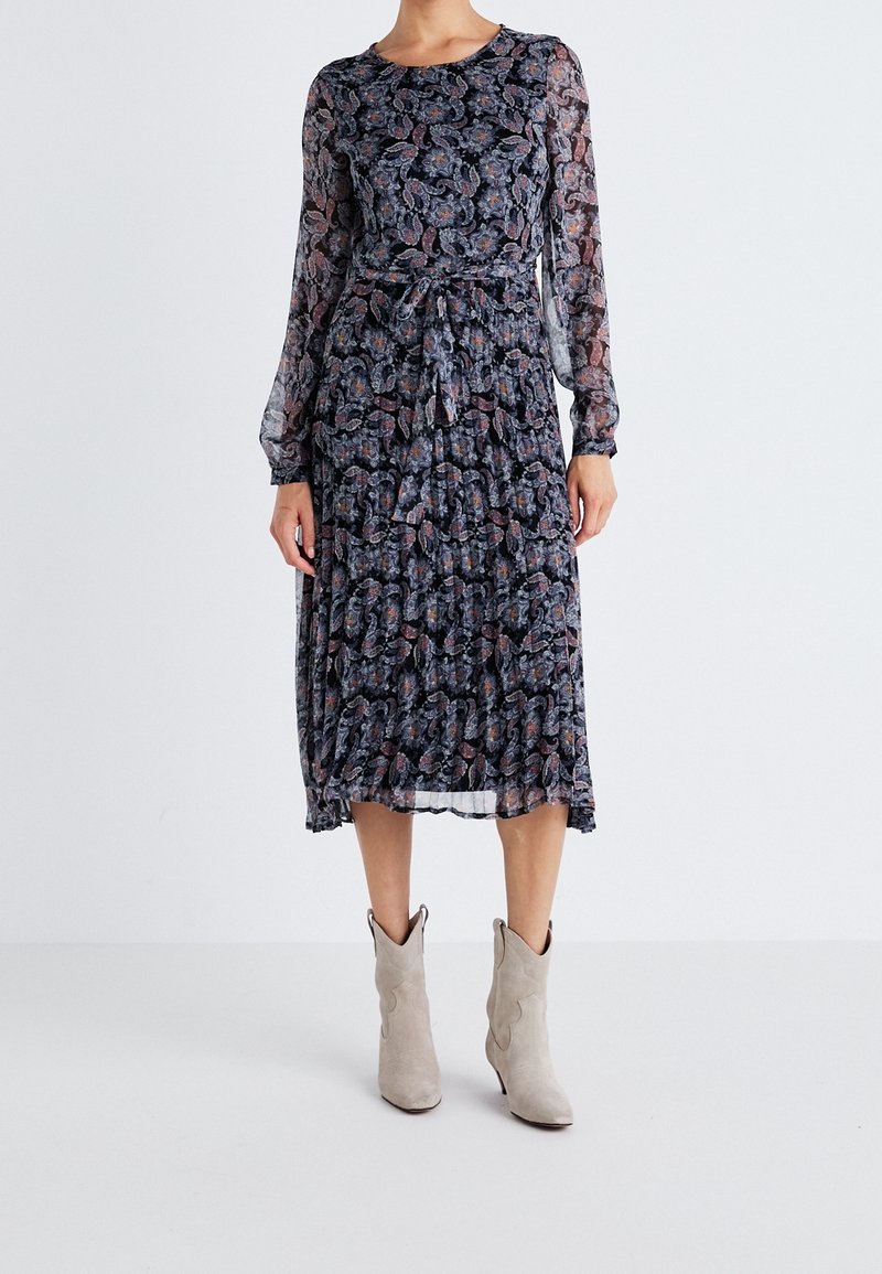 Långärmad, marinblå klänning med blommönster, midjeband och lager på lager-textur. Bärs med ljusgrå cowboystövlar.