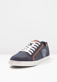 Džínové tenisky s námořnickou modrou svrškem, bílými tkaničkami a hnědými koženými detaily. Obsahují logo na patě a kontrastní šití po straně.