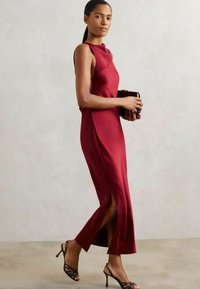 Figurbetontes burgunderfarbenes Kleid mit hohem Kragen und seitlichen Schlitzern. Model hält eine schwarze Clutch und trägt schwarze Sandalen mit Riemchen. Glattes Material.