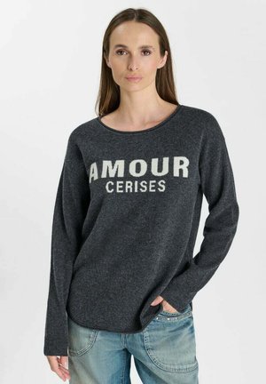 Vrouw met lang bruin haar die een donkergrijze trui met de tekst "AMOUR CERISES" en lichtblauwe spijkerbroek draagt, staand met één hand in de zak.