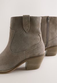 Grå ruskind ankelstøvler har en buet overkant, sidesyninger, en læderstrop til at trække i samt en lynlåslukning. Den stablede hæl har en mønstret finish.