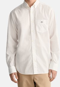 Bílá košile s dlouhým rukávem a knoflíkovým zapínáním, s kapsou na levé straně hrudníku a malým vyšitým logem. Vyrobena z hladké bavlněné látky.