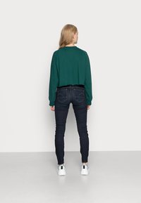 Zöld rövid fazonú pulóver hosszú ujjal, sötét, slim fit farmernadrággal párosítva. A modellen fehér sportcipő van. Minimalista háttér.