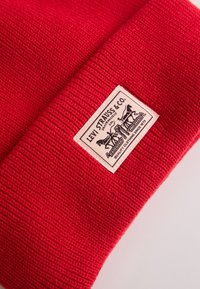 Rode gebreide muts met een omgeslagen rand en een beige Levi's label met zwarte tekst en grafisch logo. De textuur is geribbeld en glad.