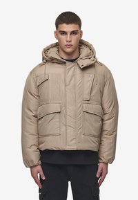 Beige pufferjakke med hette, som har flere frontlommer, glidelås og elastiske mansjetter. Glatt, polstret stoffstruktur.