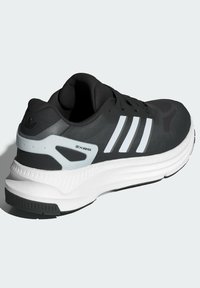 Zapatilla deportiva negra con diseño de tres rayas blancas, parte superior texturizada, suela acolchada y acentos grises. Presenta un logotipo de marca visible y una construcción robusta.