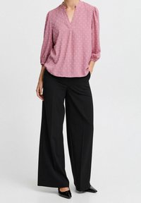 Personne portant un blouse texturée rose à manches 3/4 et un pantalon noir à jambes larges, debout avec une main dans la poche.