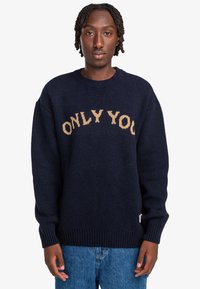 Marinblå stickad tröja med "ONLY YOU" i beige text, rundad hals, ribbade muddar och nederkant. Mjuk yta och oversized passform.