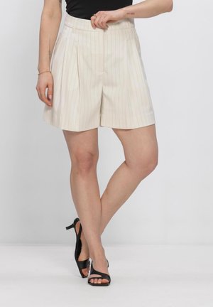 Frau trägt hoch taillierte cremefarbene Shorts mit Nadelstreifen und schwarze Sandalen mit Absatz, steht mit gekreuzten Beinen vor einem schlichten Hintergrund.