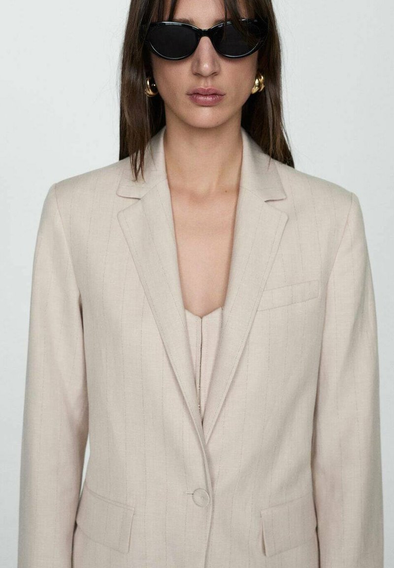 Frau trägt beigen Nadelstreifen-Blazer, passendes Oberteil, schwarze ovale Sonnenbrille und goldene Creolen vor einem schlichten Hintergrund.