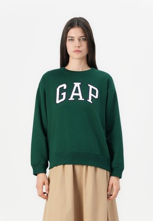 Felpa verde con lettering bianco e blu navy "GAP", scollatura a girocollo, spalle cadenti e polsini a costine, indossata sopra una gonna marrone chiaro.