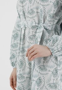 Kevyt, kuviollinen mekko pehmeässä harmaassa, jossa on vihreitä kukkakuvioita. Mekossa on sidottava vyötärö ja kootut elastiset hihansuut sopivuutta ja mukavuutta varten.