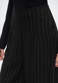 Prim-plan cu pantaloni negri cu talie înaltă, cu dungi verticale subțiri albe, purtați cu un top negru cu mâneci lungi, mâinile relaxate pe lângă corp.