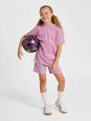 Jong meisje in roze sportkleding die een paarse en zwarte voetbal vasthoudt, staand met een lichte glimlach tegen een effen achtergrond.