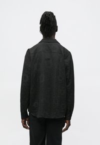 Črna srajca z zapenjanjem na gumbe in zapletenim temnim vzorcem. Ima ovratnik in dolge rokave, izdelana je iz gladke, teksturirane tkanine.