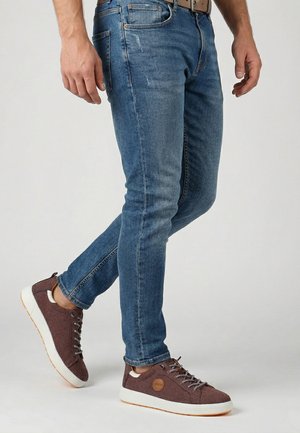 Mand iført blå slim-fit jeans og brune snørebåndssneakers stående foran en ensfarvet baggrund.