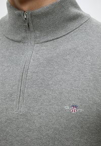 Gros plan d'un pull en maille gris avec un col à fermeture éclair quart et un petit logo bouclier brodé avec des étoiles et des rayures.