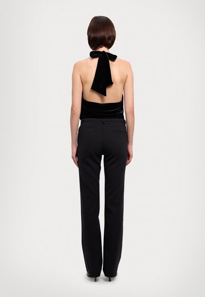 Top halter de veludo preto com costas abertas, apresentando um detalhe em laço. Combinado com calças pretas ajustadas e sapatos de salto alto.