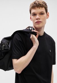 Μαύρο βαμβακερό T-shirt με κοντά μανίκια και στρογγυλή λαιμόκοψη, με ένα μικρό λογότυπο στο στήθος. Το άτομο κρατάει ένα μαύρο σακίδιο με φερμουάρ.