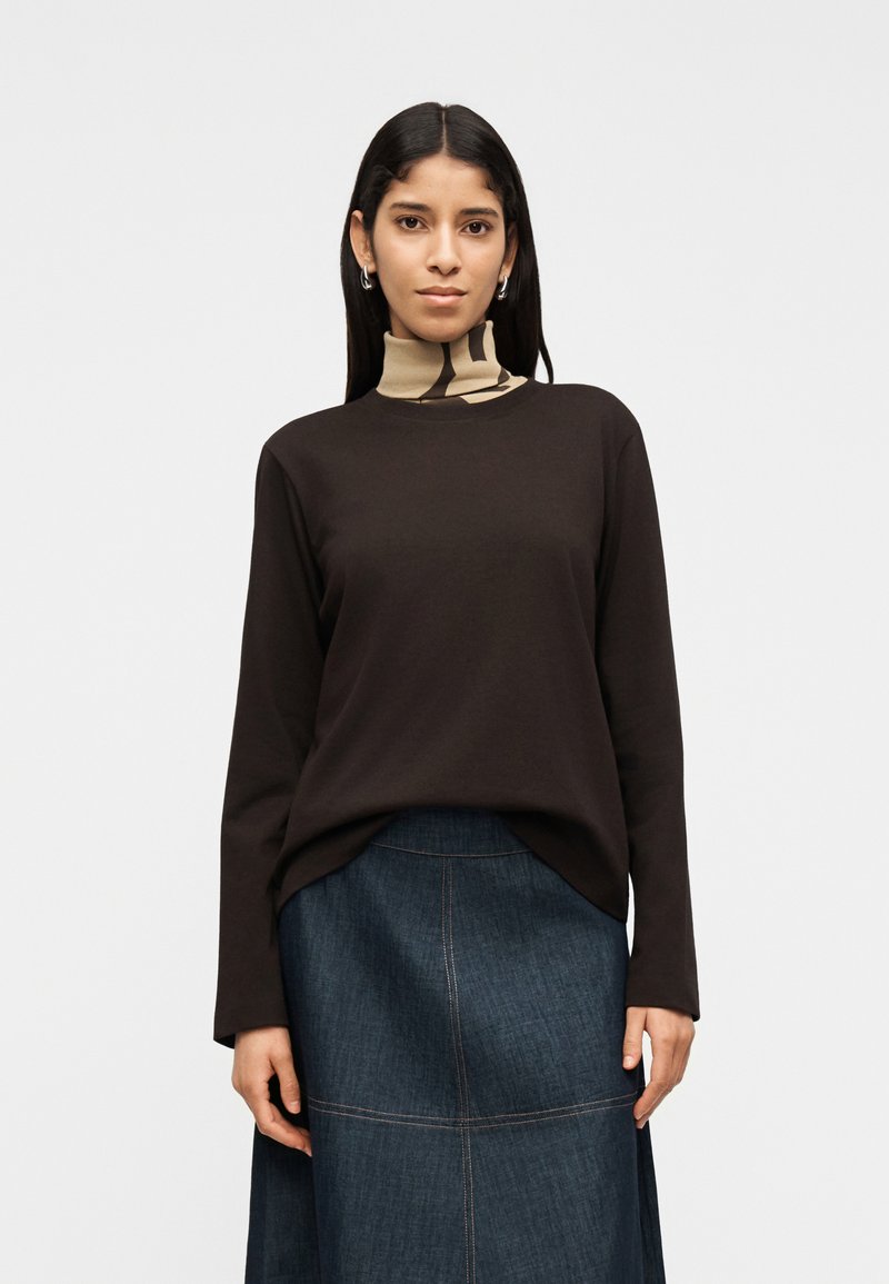 Γυναίκα με μακριά σκούρα μαλλιά που φοράει ασημένια σκουλαρίκια κρίκους, μαύρο μακρυμάνικο μπλουζάκι πάνω από μπεζ ζιβάγκο με σχέδιο και σκούρα τζιν φούστα.