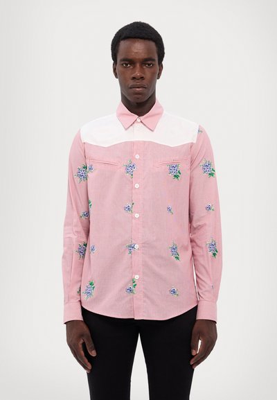 Chemise à manches longues rayée rose avec un empiècement blanc, ornée de broderies florales bleues, avec une fermeture à boutons et deux poches poitrine. Texture douce.