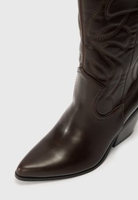 Bottine en cuir marron avec un bout pointu, un design floral cousu et un talon empilé. Texture lisse avec des coutures marron contrastantes.