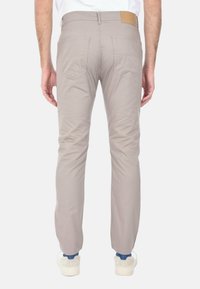 Pantaloni beige slim-fit con tasche posteriori e una toppa in pelle marrone alla vita, mostrati da dietro su una persona in piedi.