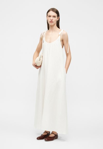 Γυναίκα που στέκεται, φοράει ένα μακρύ λευκό αμάνικο φόρεμα με δέσιμο στους ιμάντες, κρατάει μια μπεζ τσάντα χειρός και καφέ φλατ παπούτσια με ιμάντες στον αστράγαλο.