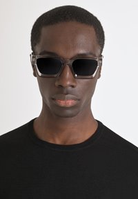 Occhiali da sole neri con una montatura trasparente e angolare e lenti scure. Indossati da una persona in un maglione nero a girocollo su uno sfondo chiaro.