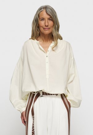 Blusa crema a maniche lunghe con placket abbottonato, colletto arricciato e polsini oversize; abbinata a pantaloni bianchi e una cintura marrone.