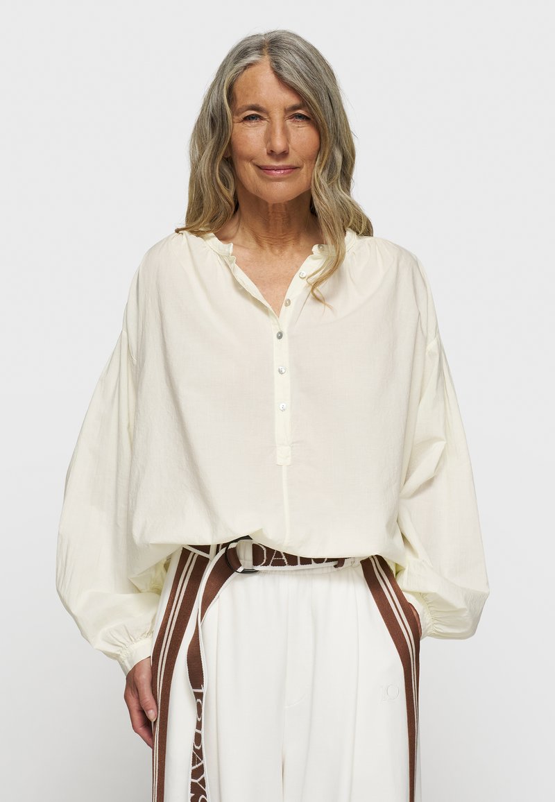 Blusa crema a maniche lunghe con placket abbottonato, colletto arricciato e polsini oversize; abbinata a pantaloni bianchi e una cintura marrone.