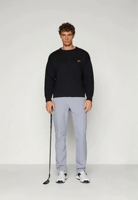 Svart strikket genser med ribbestrikkede mansjetter, grå skreddersydde bukser og svarte og hvite golfsko, holder en golfklubbe.