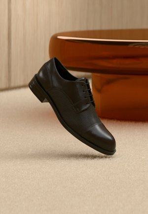 Scarpa elegante da uomo in pelle nera con pannelli laterali traforati, sospesa sopra un pavimento beige coperto da moquette accanto a un tavolo rotondo marrone lucido.